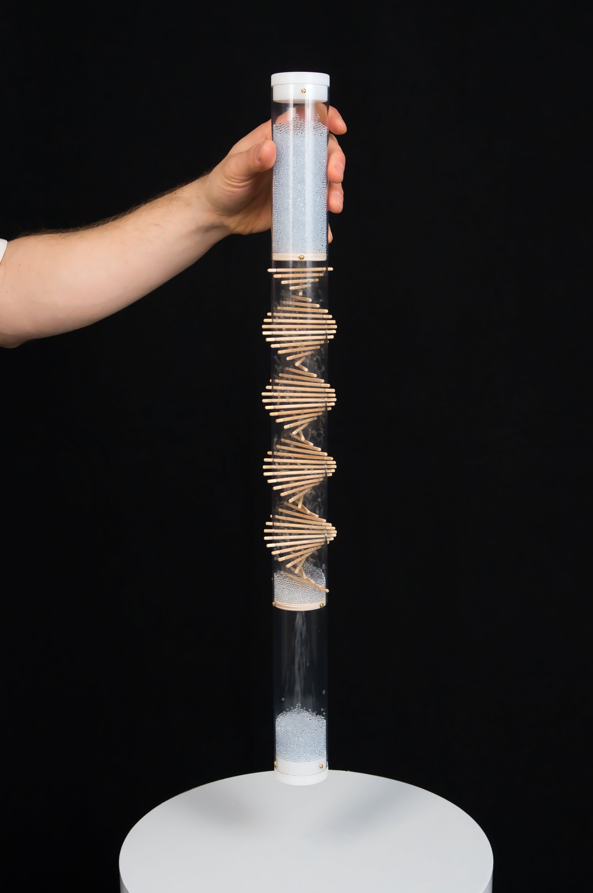 Person holding a small spiral sounds rainstick by Rainstick Dreams. The rainstick is comprised of a clear tube with a wooden spiral helix going through it. Pale blue glass beads flow down. The rainstick is being held up above a white pedestal, with a black backdrop.