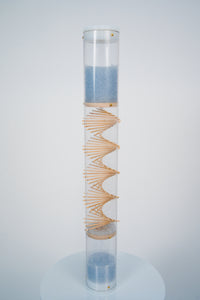 A clear spiral sounds rainstick by Rainstick Dreams has blue glass beads flowing from top to bottom through the wooden spiral helix. The top and bottom of the clear tube is enclosed with white end caps. The rainstick is standing on a white pedestal against a white background.