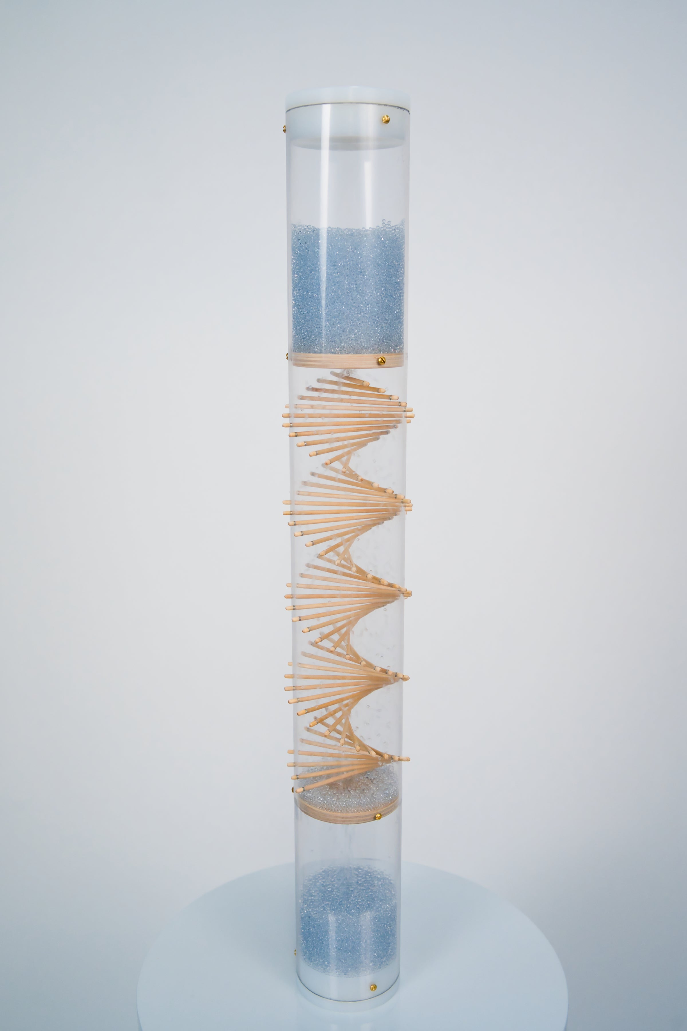 A clear spiral sounds rainstick by Rainstick Dreams has blue glass beads flowing from top to bottom through the wooden spiral helix. The top and bottom of the clear tube is enclosed with white end caps. The rainstick is standing on a white pedestal against a white background.