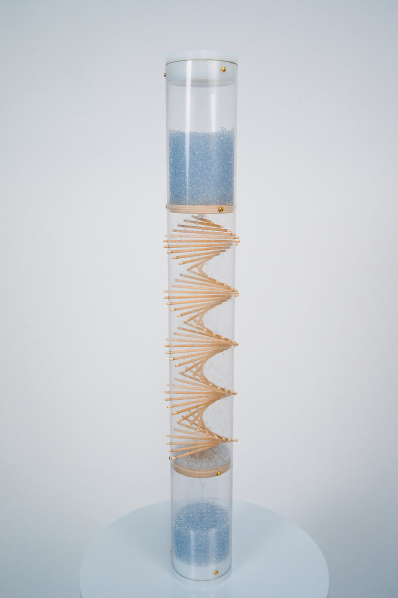 A clear spiral sounds rainstick by Rainstick Dreams has blue glass beads flowing from top to bottom through the wooden spiral helix. The top and bottom of the clear tube is enclosed with white end caps. The rainstick is standing on a white pedestal against a white background.