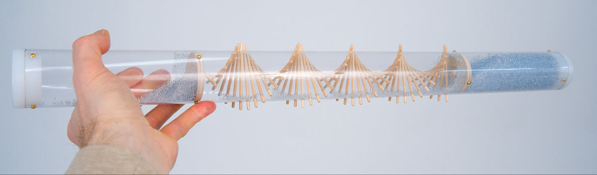 An outstretched hand holds a clear spiral rainstick horizontally against a white background. The visually stunning rainstick contains pale blue glass beads. White caps enclose the long duration rainstick sound healing instrument on both ends.