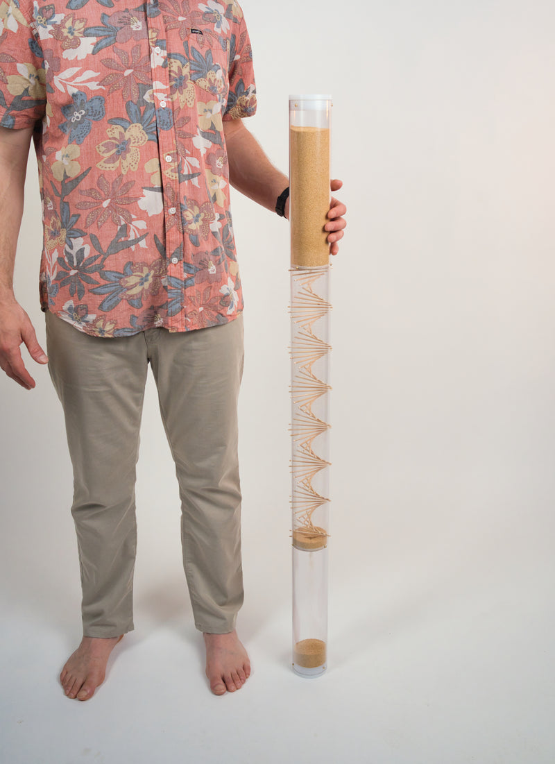Person standing next to a 4' tall amaranth rainstick instrument with a wooden spiral going through a clear tube. The instrument makes a gentle rain sound for 10 minutes and the person is cradling the top of it in their hand.