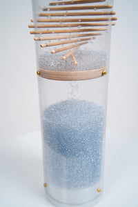 The bottom of a rainstick housed in a clear tube with a white end cap enclosing it. Pale blue glass beads reflect light as they descend down wooden sticks protruding through the clear tube in a spiral pattern. The beads fall and collect at the bottom. The beads produce a sound as they collide with the sticks and the tube, making this a long duration instrument. It’s against a white background.