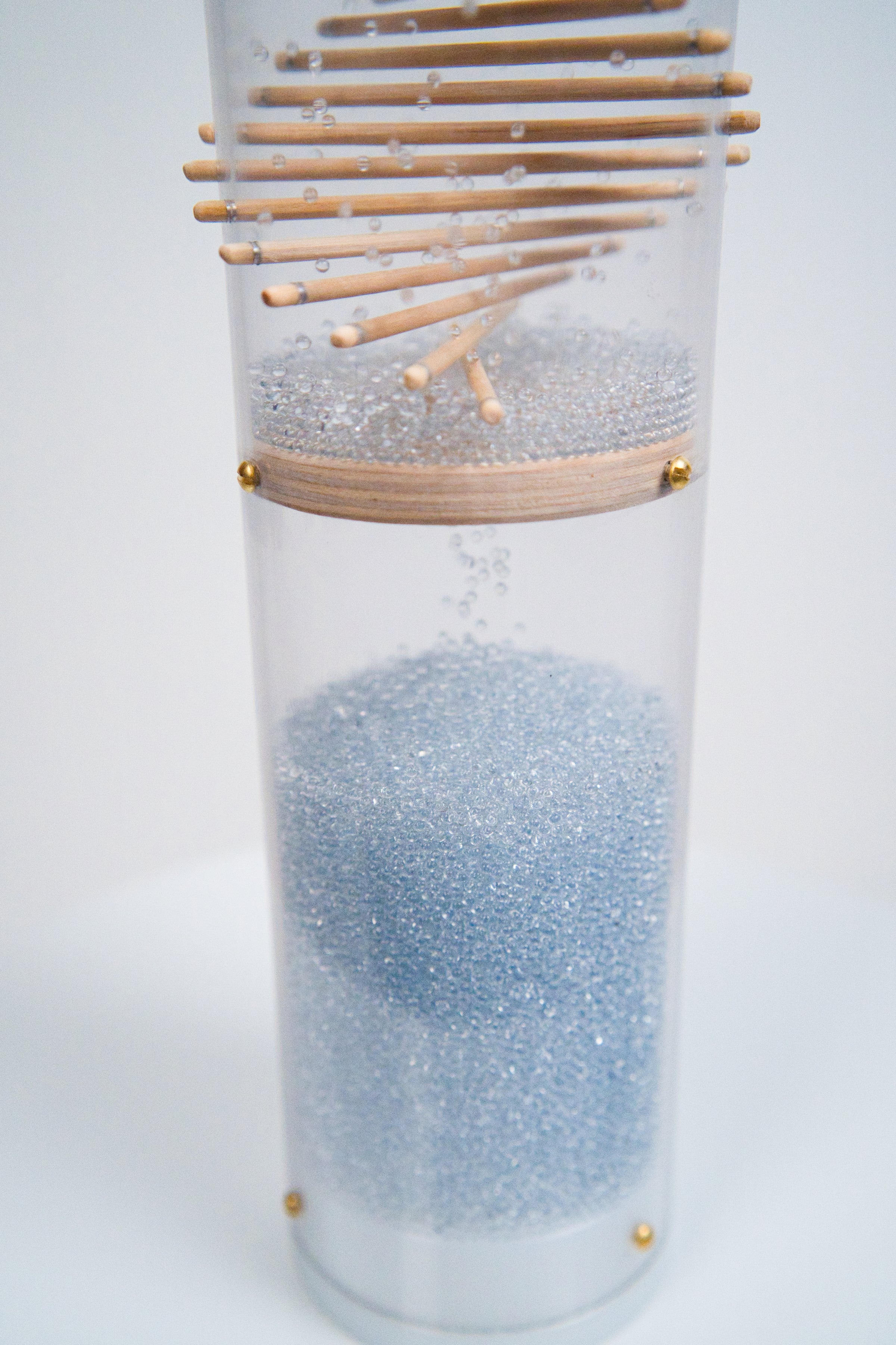 The bottom of a rainstick housed in a clear tube with a white end cap enclosing it. Pale blue glass beads reflect light as they descend down wooden sticks protruding through the clear tube in a spiral pattern. The beads fall and collect at the bottom. The beads produce a sound as they collide with the sticks and the tube, making this a long duration instrument. It’s against a white background.