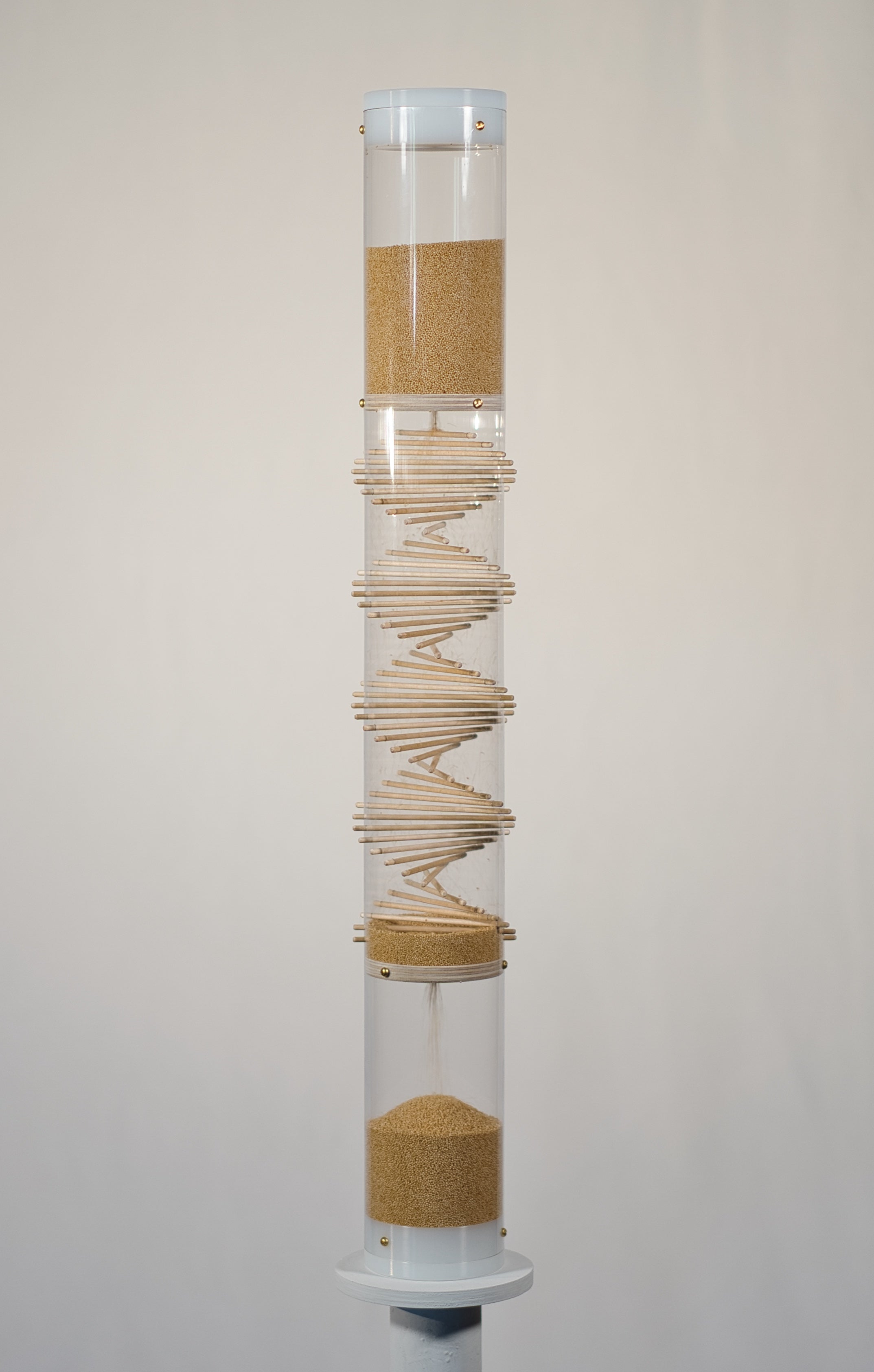 3.5 minute handcrafted spiral sounds rainstick displayed on a pedestal. The rainstick is 2 feet tall and has a clear tube. The amaranth is a tan seed. The end caps of the instrument are white.