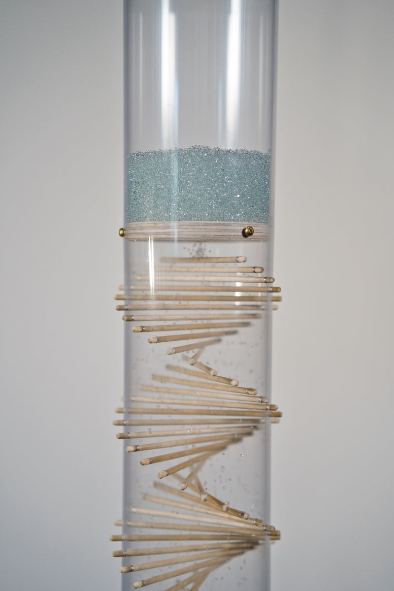 Blue glass microbeads fall inside a clear rainstick tube that features a visible wooden spiral. 