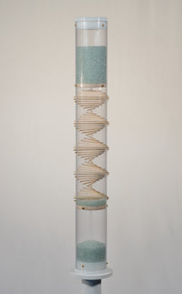 A spiral sounds midway microbeads rainstick stands 2 feet tall, and is displayed on a pedestal against a white background. The glass microbeads are blue and sparkle in the light. The rainstick tube is clear, and there is a visible wooden spiral down the middle of the instrument.