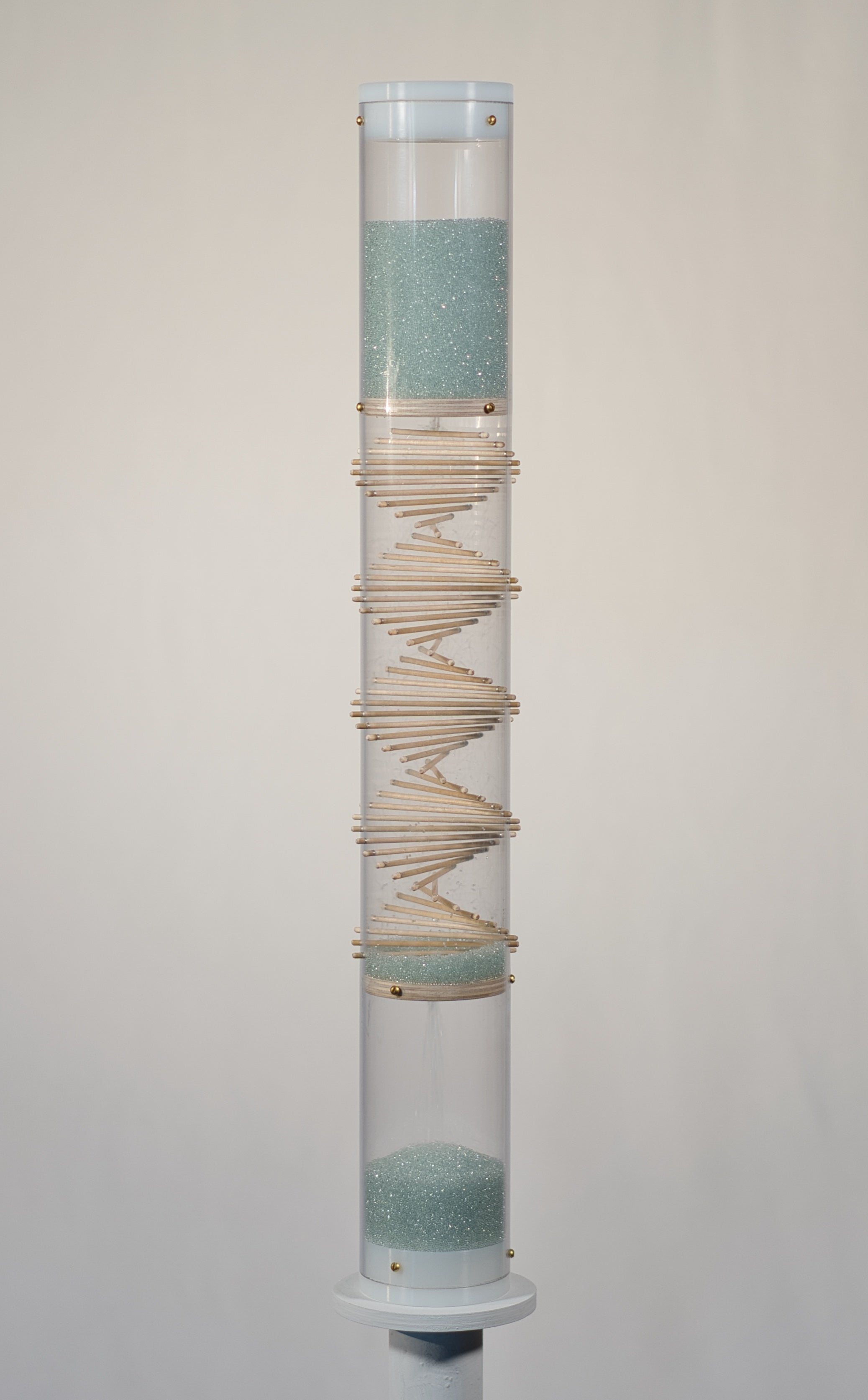 A spiral sounds midway microbeads rainstick stands 2 feet tall, and is displayed on a pedestal against a white background. The glass microbeads are blue and sparkle in the light. The rainstick tube is clear, and there is a visible wooden spiral down the middle of the instrument.