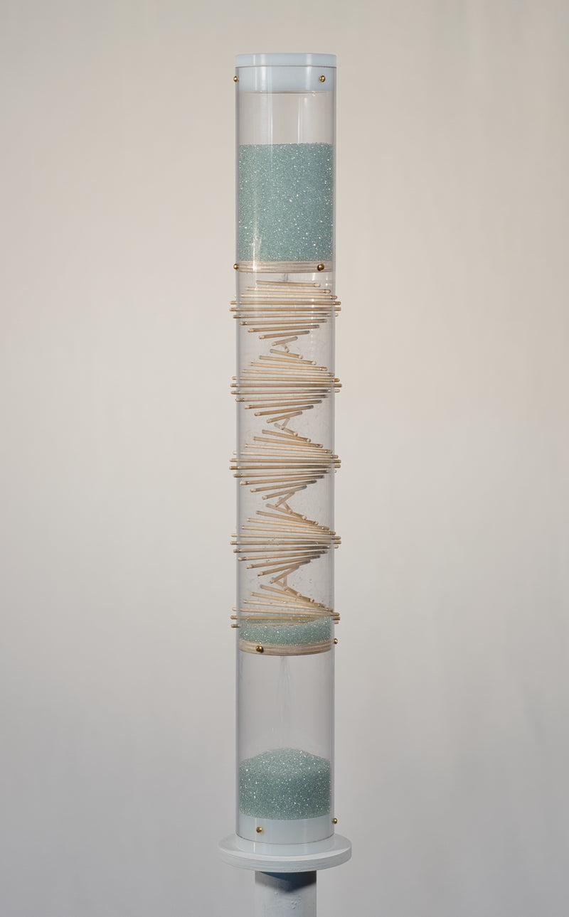 A spiral sounds midway microbeads rainstick stands 2 feet tall, and is displayed on a pedestal against a white background. The glass microbeads are blue and sparkle in the light. The rainstick tube is clear, and there is a visible wooden spiral down the middle of the instrument.