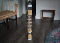 A 2' tall clear spiral sounds rainstick filled with amaranth, standing on the floor of a home. There is furniture nearby. This gentle sounding rainstick instrument is often paired with other instruments for sound baths, sound healing, and reiki.