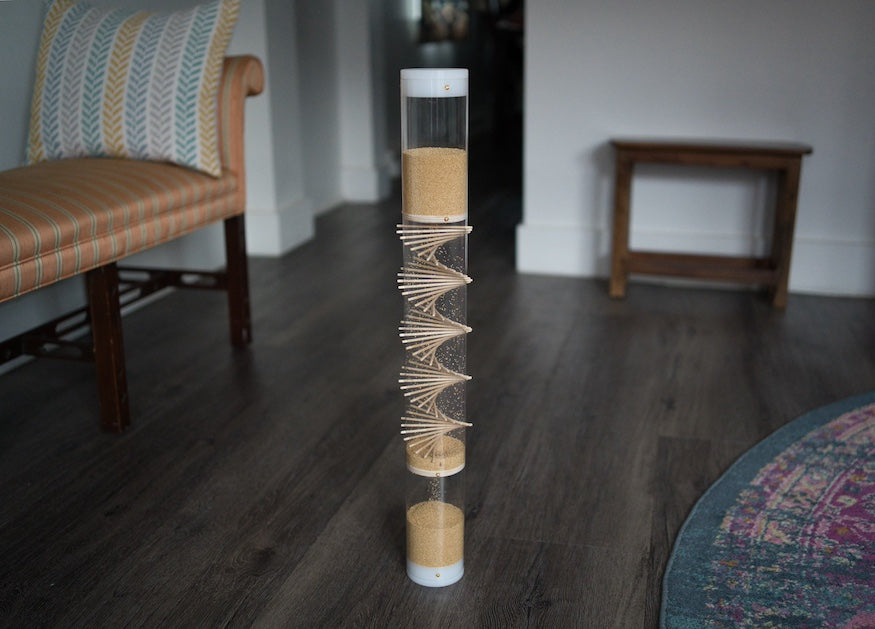 A 2' tall clear spiral sounds rainstick filled with amaranth, standing on the floor of a home. There is furniture nearby. This gentle sounding rainstick instrument is often paired with other instruments for sound baths, sound healing, and reiki.