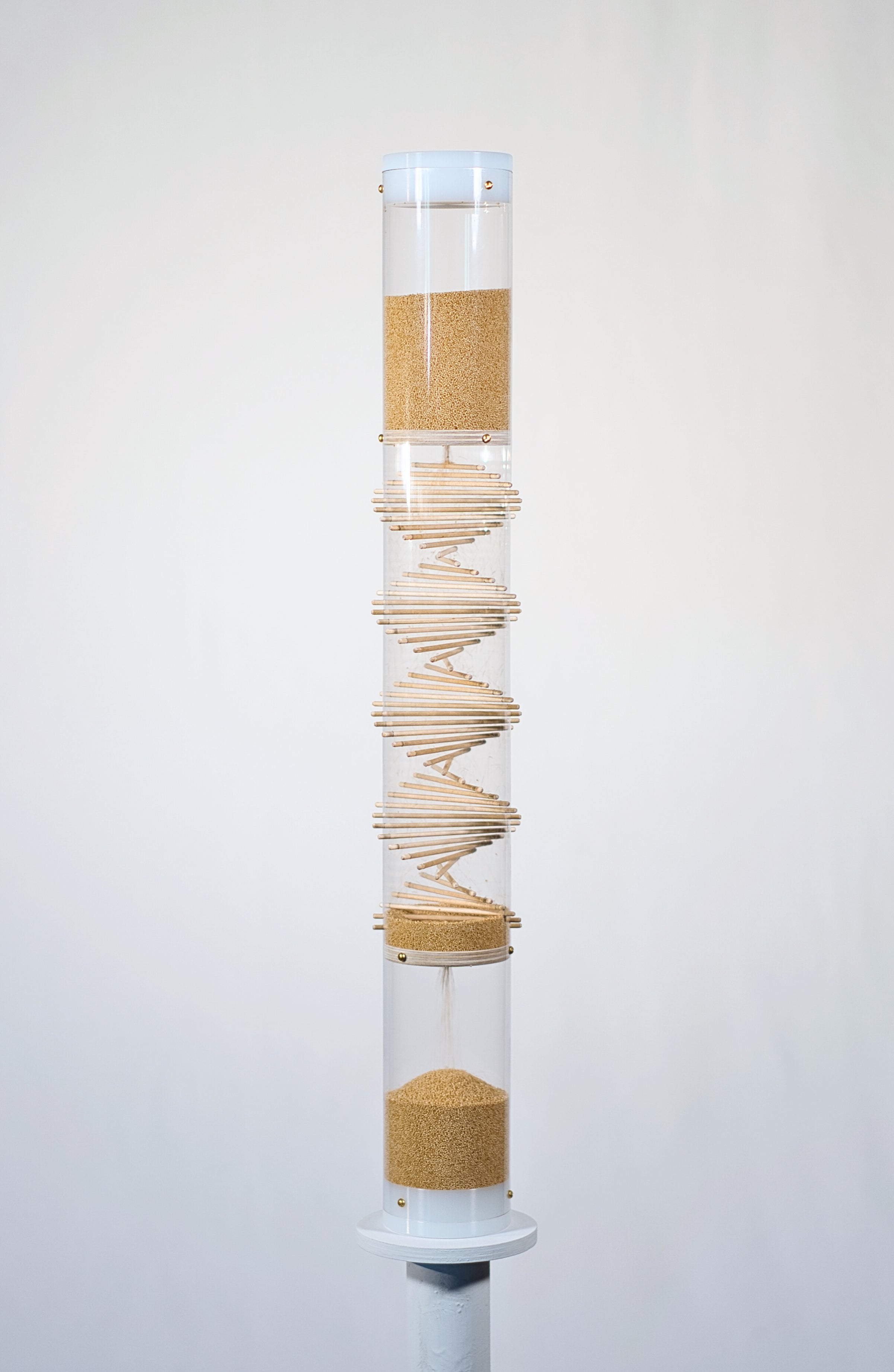 Cylindrical clear tube stands 2’ tall on a white circular pedestal against a white background. The tube has wooden sticks going through it in a spiral pattern, and tan amaranth seed is flowing from the top down to the bottom. The amaranth seeds bounce among the sticks and against the tube and create a gentle rain sound for 3.5 minutes as they descend down the spiral sounds rainstick by Rainstick Dreams.