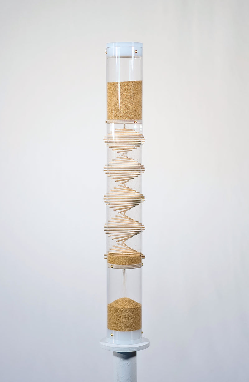 Cylindrical clear tube stands 2’ tall on a white circular pedestal against a white background. The tube has wooden sticks going through it in a spiral pattern, and tan amaranth seed is flowing from the top down to the bottom. The amaranth seeds bounce among the sticks and against the tube and create a gentle rain sound for 3.5 minutes as they descend down the spiral sounds rainstick by Rainstick Dreams.