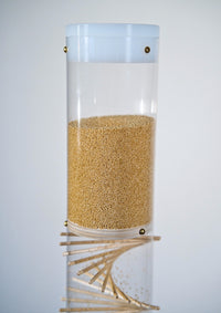 A clear cylinder enclosed with a solid white cap on top contains tiny amaranth seeds, a wooden funnel disk, and a wooden spiral pattern. It is a 3.5 minute long duration spiral sounds rainstick by Rainstick Dreams. It’s gentle rain sound is calming and can help relax the nervous system - just flip and listen.