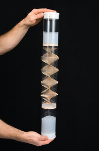 A person is holding a long duration rainstick housed in a clear tube at the top and bottom, where the white end caps enclose the cylindrical instrument. A wooden spiral protrudes through the clear tube and pale blue glass beads descend and produce a sound as they ricochet among the sticks and the tube. There’s a black background.