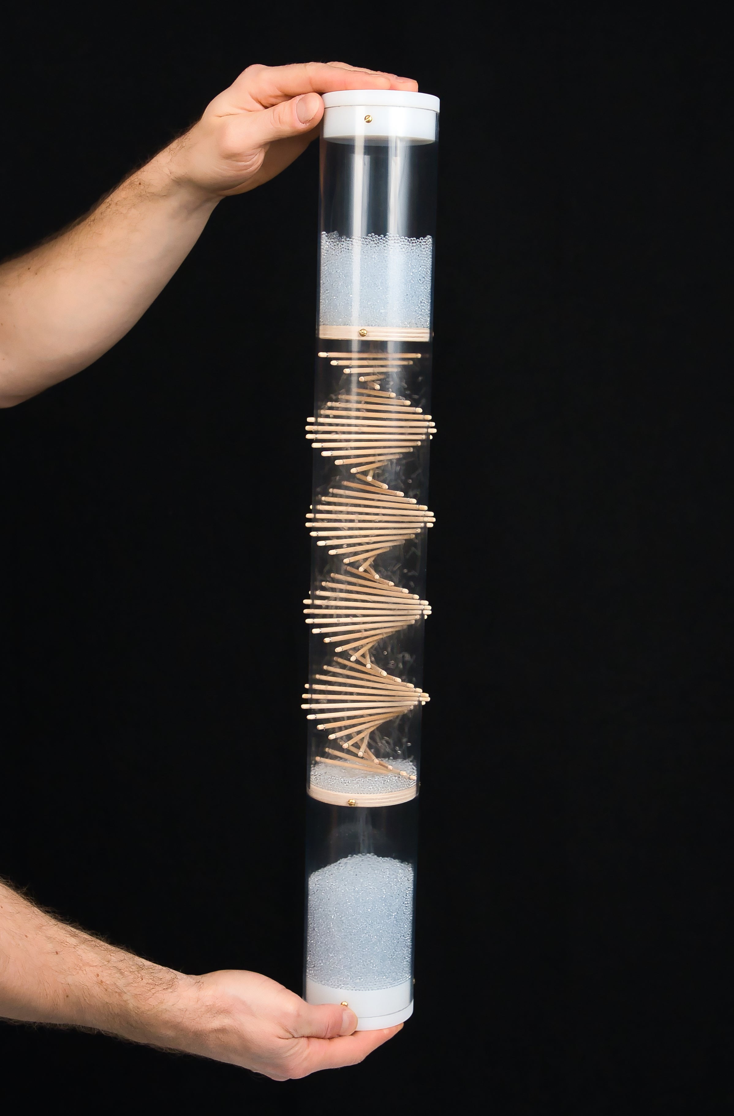 A person is holding a long duration rainstick housed in a clear tube at the top and bottom, where the white end caps enclose the cylindrical instrument. A wooden spiral protrudes through the clear tube and pale blue glass beads descend and produce a sound as they ricochet among the sticks and the tube. There’s a black background.