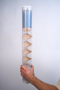 A person is holding a Midway Spiral Sounds Rainstick by Rainstick Dreams in their hand. A wooden spiral goes through the instrument, and blue glass beads flow down within the clear rainstick tube. The long duration sound healing instrument is enclosed with a white cap at the top and bottom.