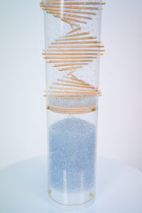 The lower portion of the Midway spiral rainstick by Rainstick Dreams. Pale blue glass beads ricochet around the wooden spiral patterned sticks that protrude through a clear tube. The beads produce sound as they fall and collide with the sticks and the tube. A white end cap encloses the bottom of the long duration Rainstick instrument.