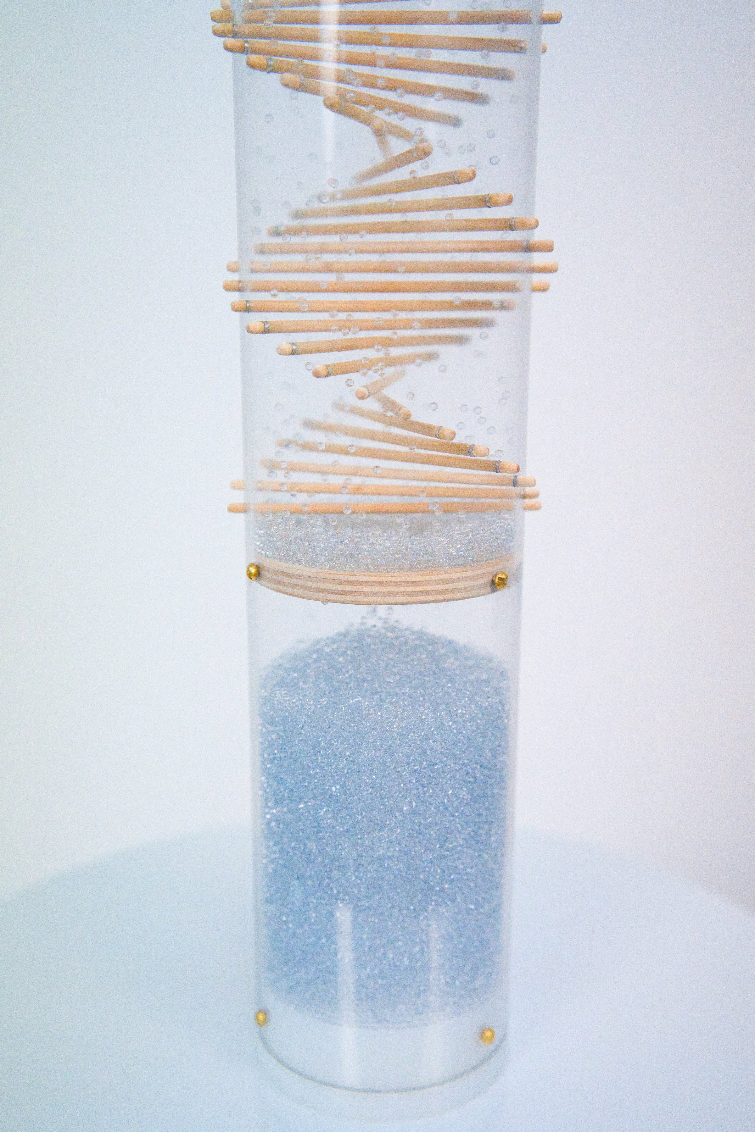 The lower portion of the Midway spiral rainstick by Rainstick Dreams. Pale blue glass beads ricochet around the wooden spiral patterned sticks that protrude through a clear tube. The beads produce sound as they fall and collide with the sticks and the tube. A white end cap encloses the bottom of the long duration Rainstick instrument.