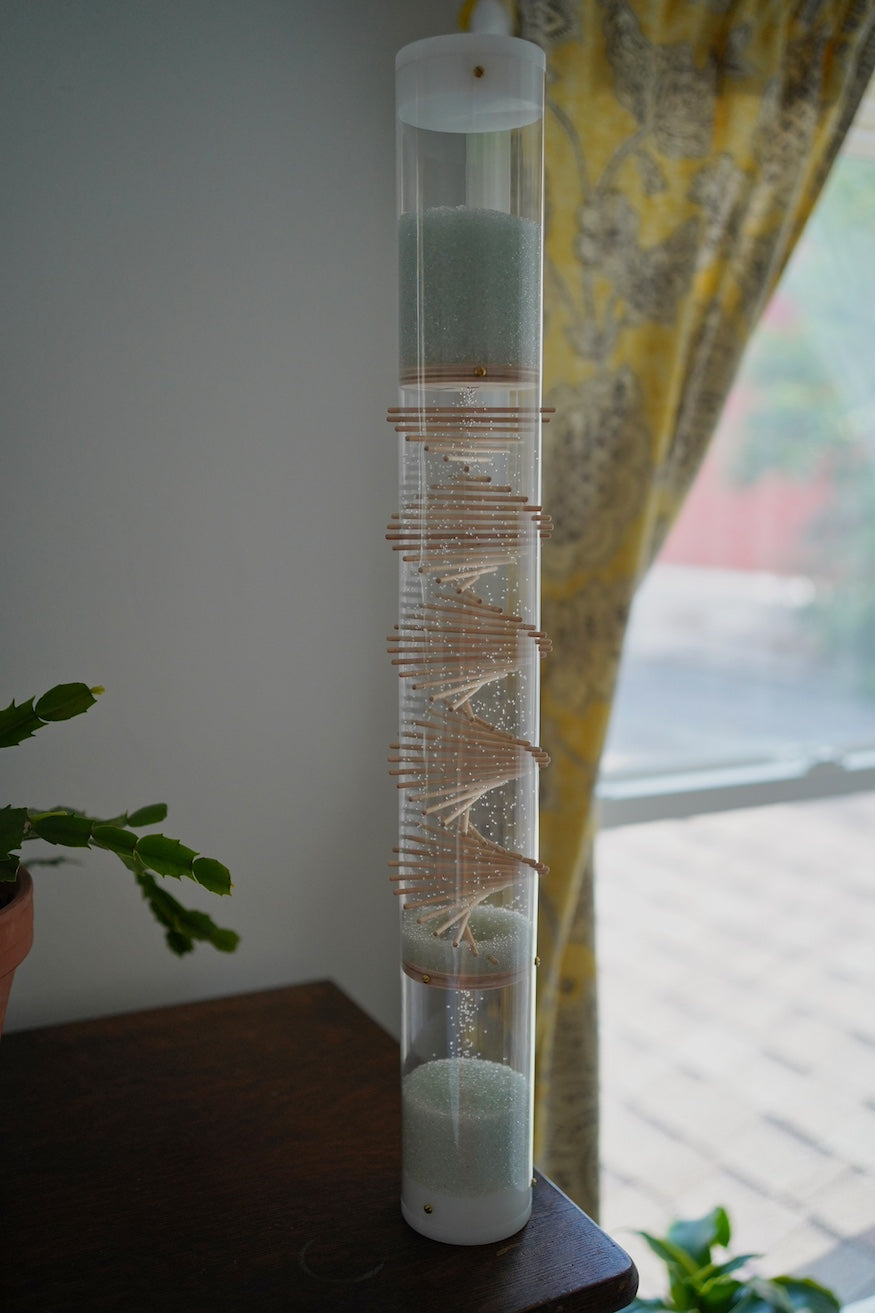 A 2' tall rainstick stands on furniture with a window and plants behind it. There is a visible wooden spiral in the clear tube, and the natural blue glass microbeads are falling inside the rainstick. This midway Microbeads Rainstick lasts for 3 minutes.