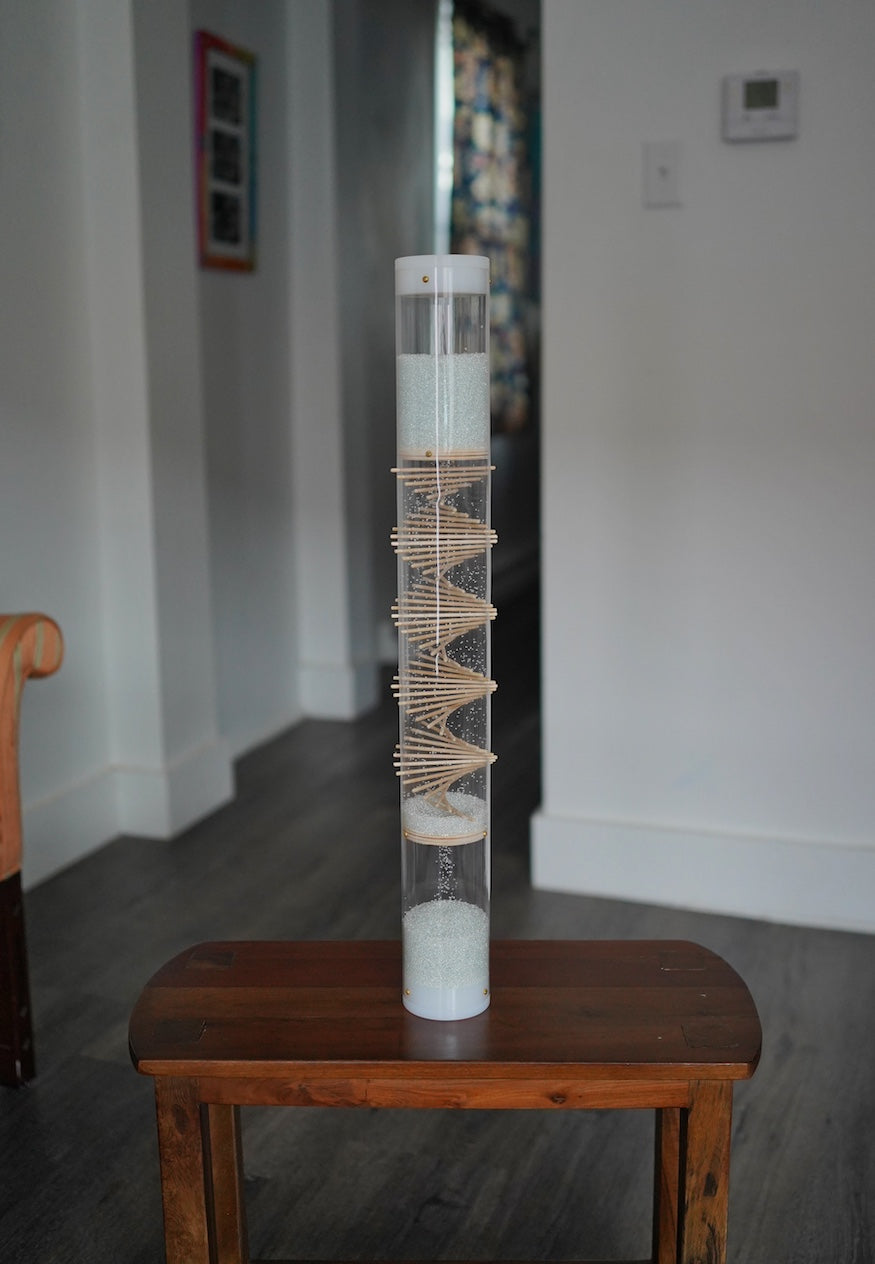 A 2 foot tall rainstick stands on a wooden table in a home. The rainstick tube is clear and has white end caps. There is a visible wooden spiral helix going through the middle portion of the tube. This is 3 minute spiral rainstick made by Rainstick Dreams.