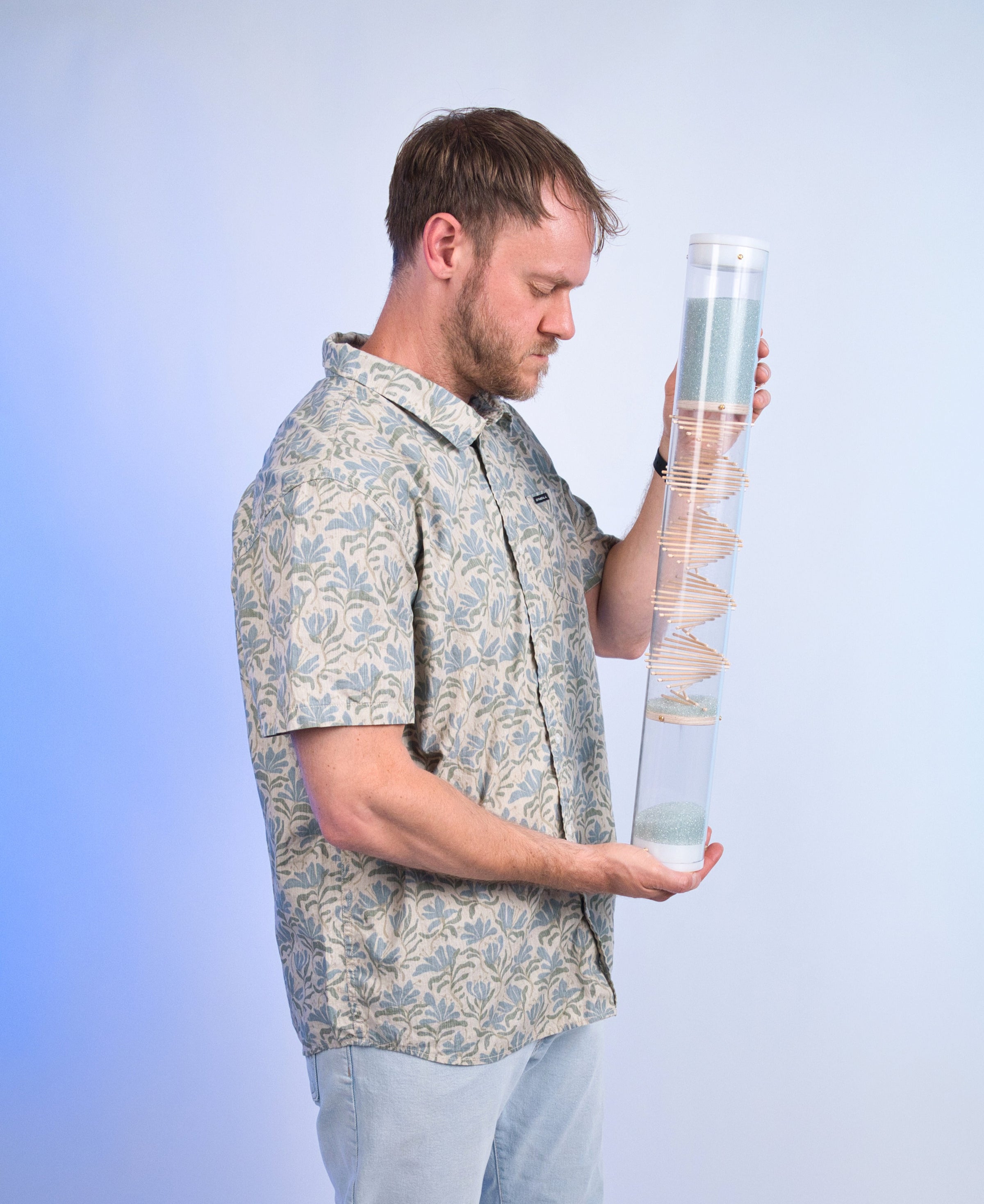 Midway 3-minute handmade glass microbeads rainstick with the visible spiral design of sticks through a clear tube, held upright by a person against a blue background