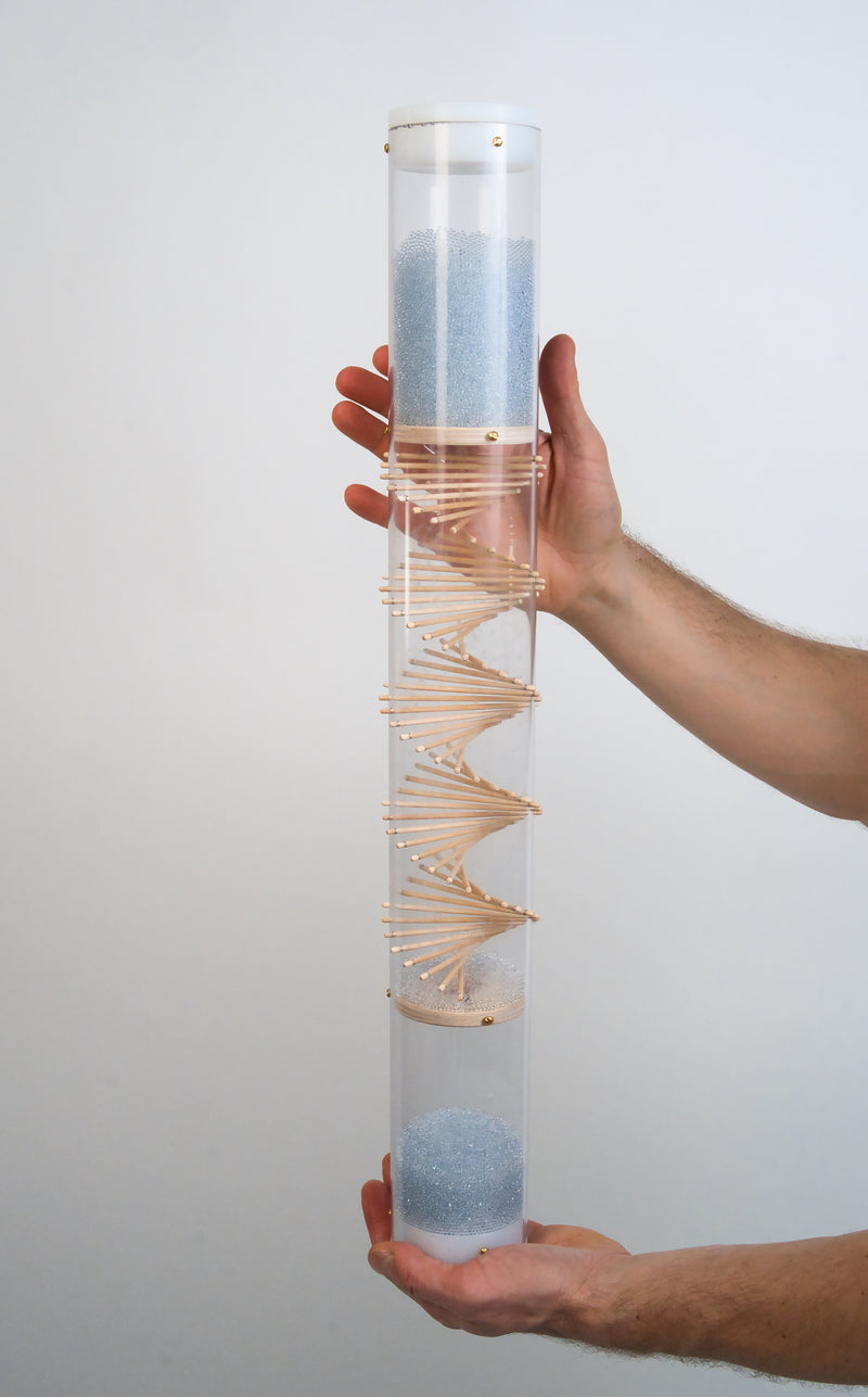 A person is holding a clear spiral sounds rainstick by Rainstick Dreams in their hands. Their hands are placed around the top and bottom white end caps of the long duration instrument. A wooden spiral helix goes through the clear Rainstick tube, and blue glass beads flow from top to the bottom.