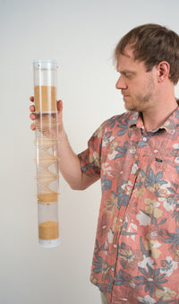 Midway 3.5-minute handmade amaranth rainstick with the visible spiral design of sticks through a clear tube, held vertically to show warm golden beads cascading inside