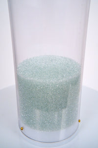 The closeup image shows a collection of shimmering pale blue glass microbeads at the bottom of a clear tube that is sealed by a white plastic end cap. The base of the medium rain sound glass beads rainstick is on a white pedestal against a white background.