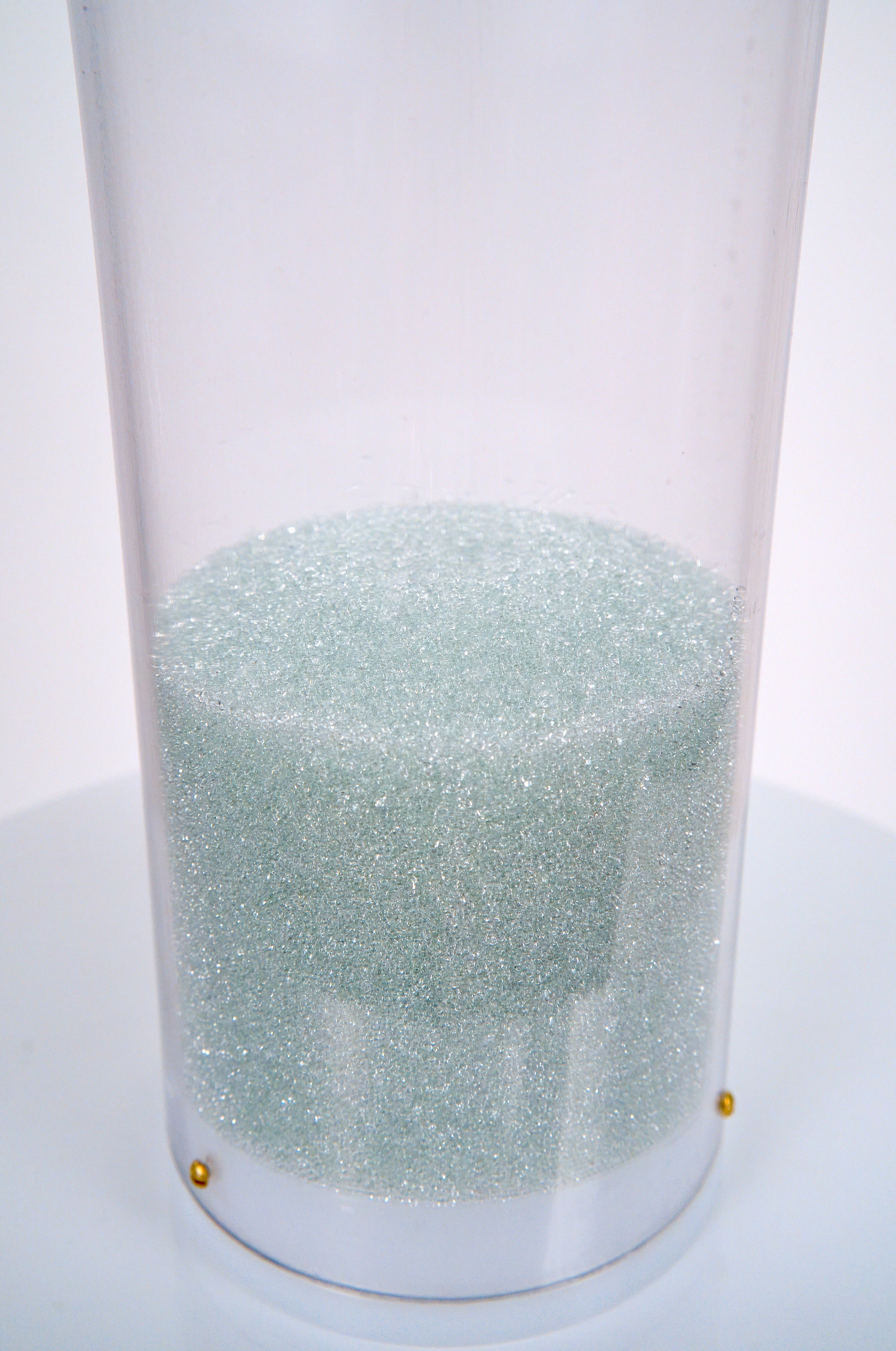 The closeup image shows a collection of shimmering pale blue glass microbeads at the bottom of a clear tube that is sealed by a white plastic end cap. The base of the medium rain sound glass beads rainstick is on a white pedestal against a white background.