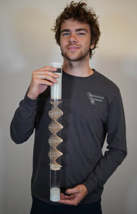 A person wearing a dark grey shirt holds a 2' tall clear rainstick in front of them. The rainstick tube is clear and its wooden spiral contrasts well against the dark grey shirt.