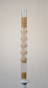 Two foot tall clear spiral sounds rainstick filled with tan amaranth seed. There is a visible wooden spiral going through the middle of the clear tube. The rainstick has white end caps on the top and bottom of the instrument. The rainstick instrument is on a white pedestal with a white background.