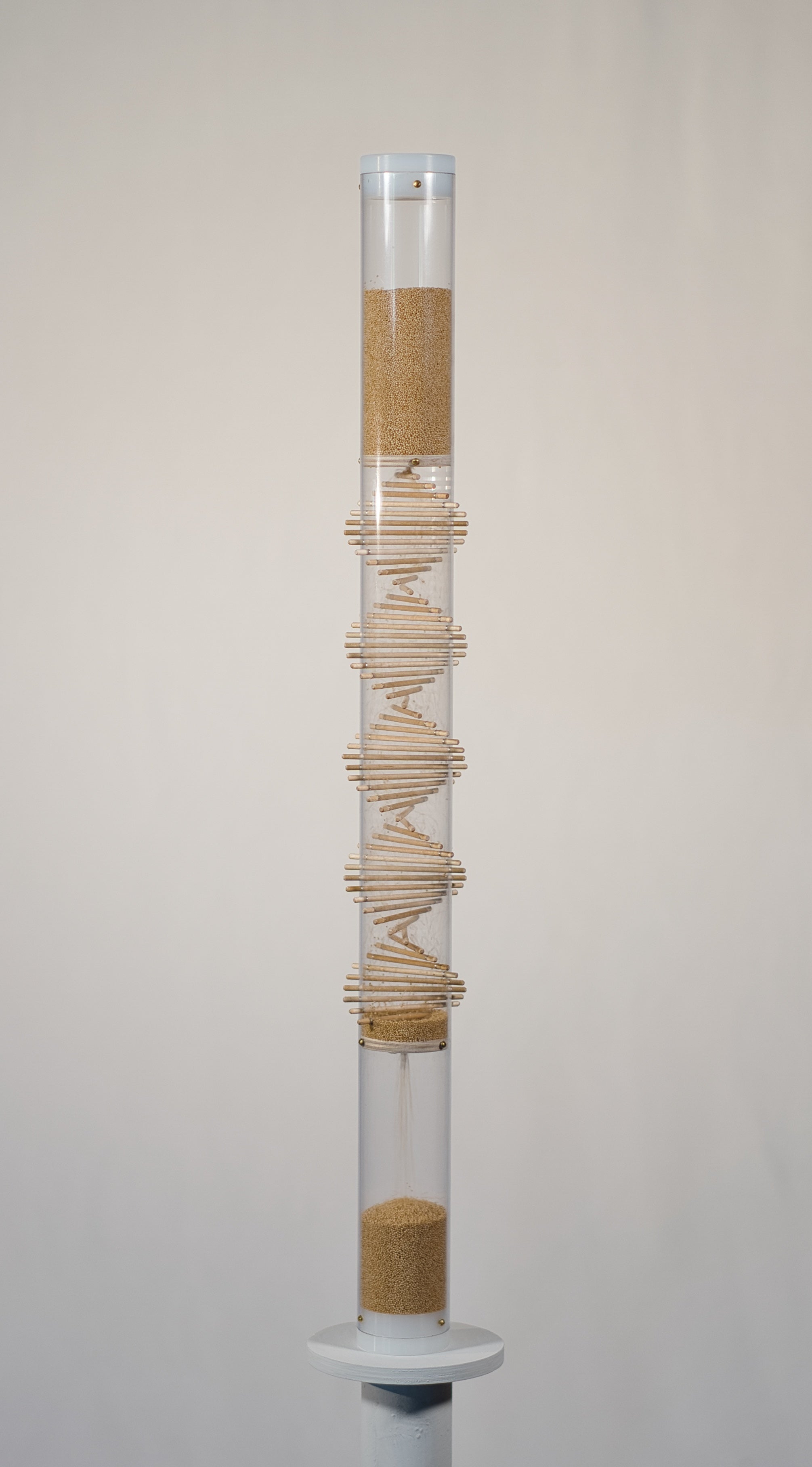 Two foot tall clear spiral sounds rainstick filled with tan amaranth seed. There is a visible wooden spiral going through the middle of the clear tube. The rainstick has white end caps on the top and bottom of the instrument. The rainstick instrument is on a white pedestal with a white background.