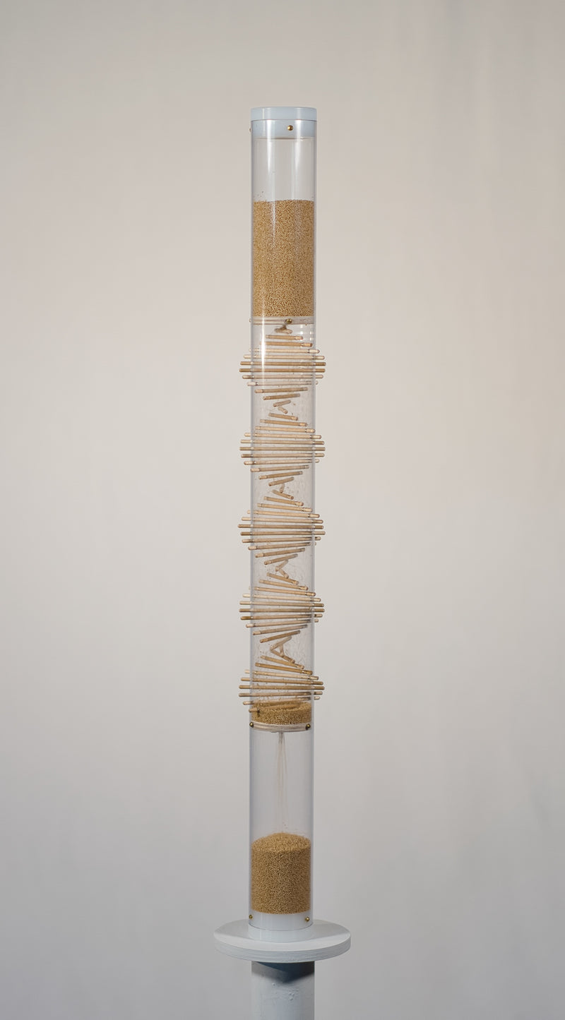 Two foot tall clear spiral sounds rainstick filled with tan amaranth seed. There is a visible wooden spiral going through the middle of the clear tube. The rainstick has white end caps on the top and bottom of the instrument. The rainstick instrument is on a white pedestal with a white background.