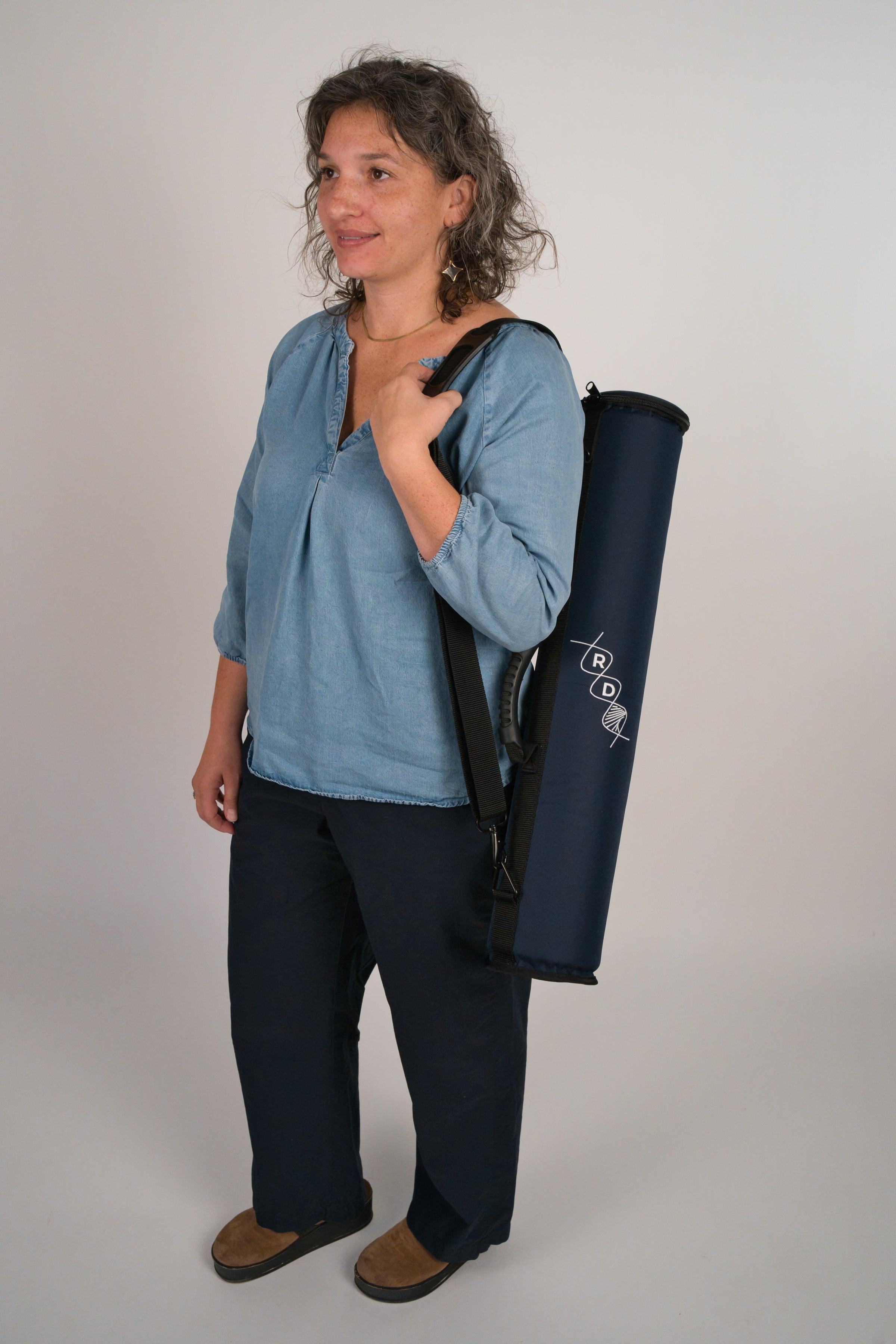 Person holding a navy blue instrument case featuring the RD helix logo over their shoulder in front of a plain background. The person is stepping forward, to the left of the camera, looking ahead, and smiling.