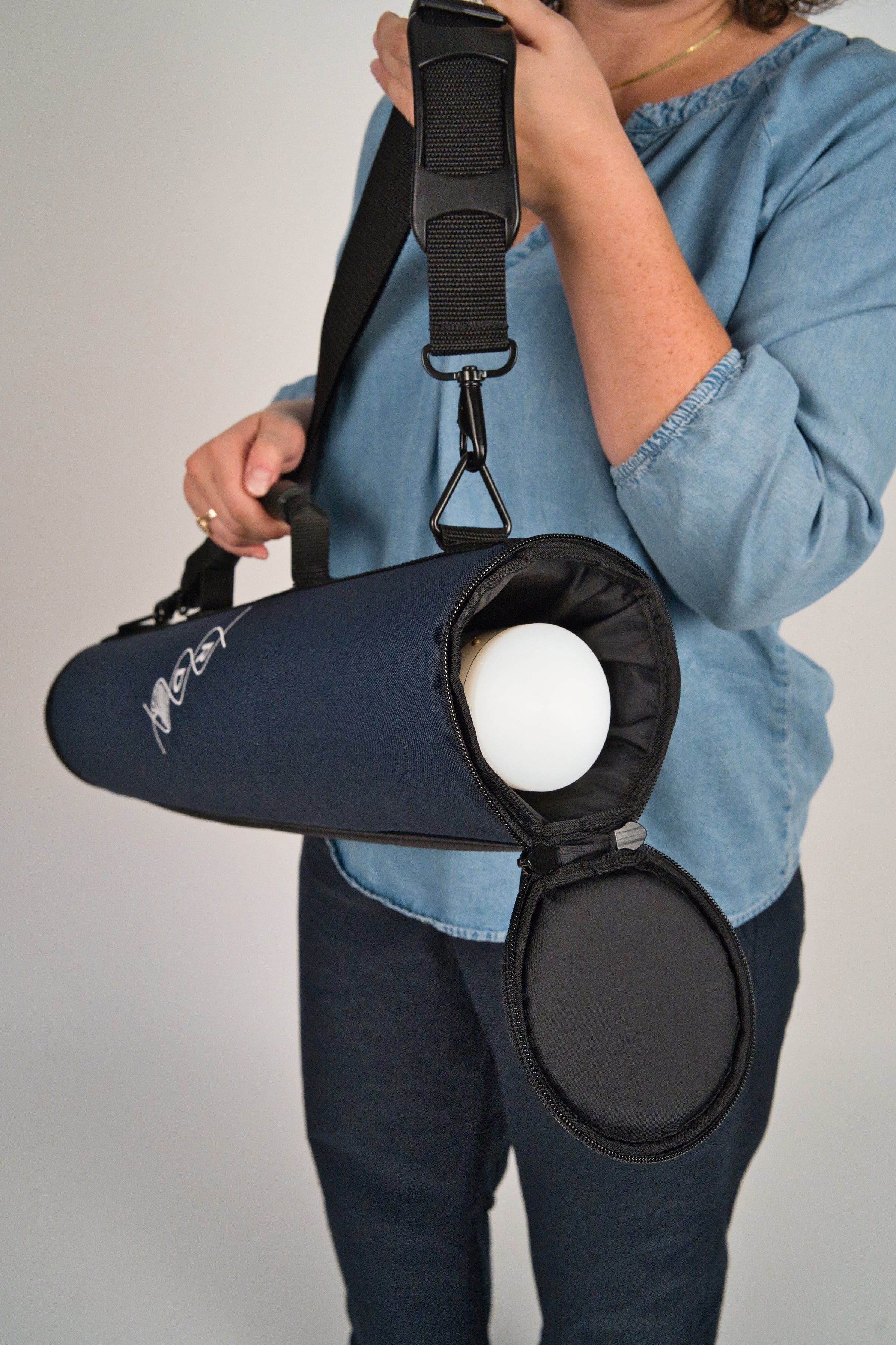 Person holding a navy blue rainstick instrument case by the strap and handle while the zipper around the top is open. The white cap of the rainstick inside the case is visible.