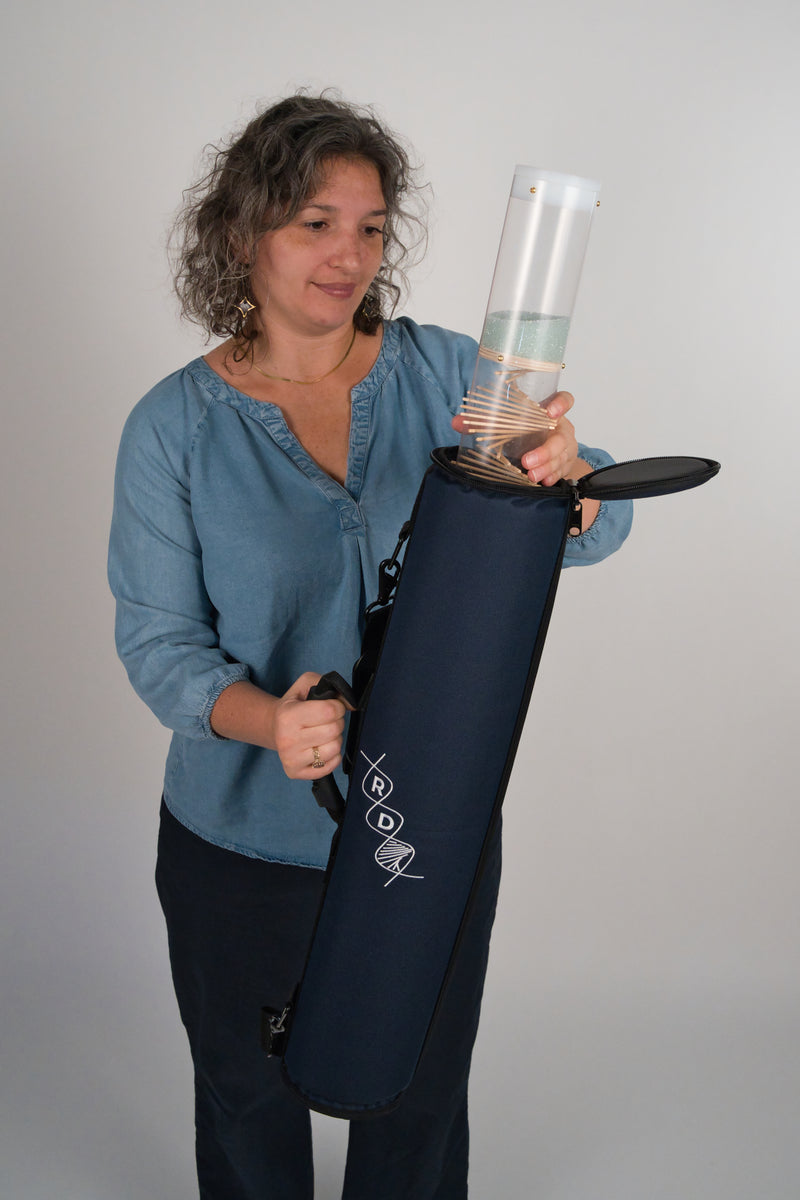 Person holding the upper portion of a clear visible spiral rainstick that is partially inside a navy blue instrument case. The person is holding the instrument case with their right hand and the rainstick with their left hand. The person is looking at the rainstick with a gentle smile on their face.