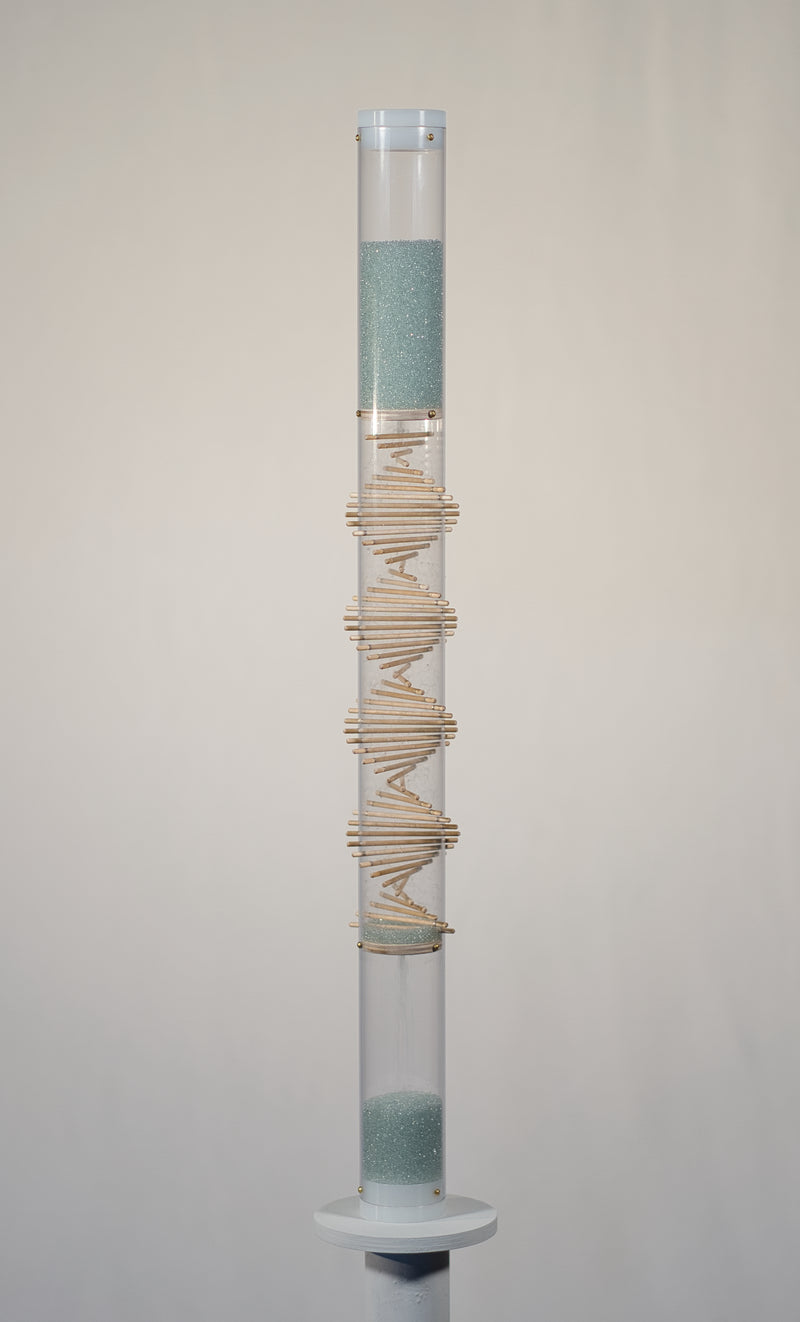 A 2 foot tall clear rainstick with a wooden spiral going down the middle has blue glass microbeadss in the top and bottom. There are white end caps on the top and bottom of the instrument. The rainstick is on a white pedestal, in front of a white background.