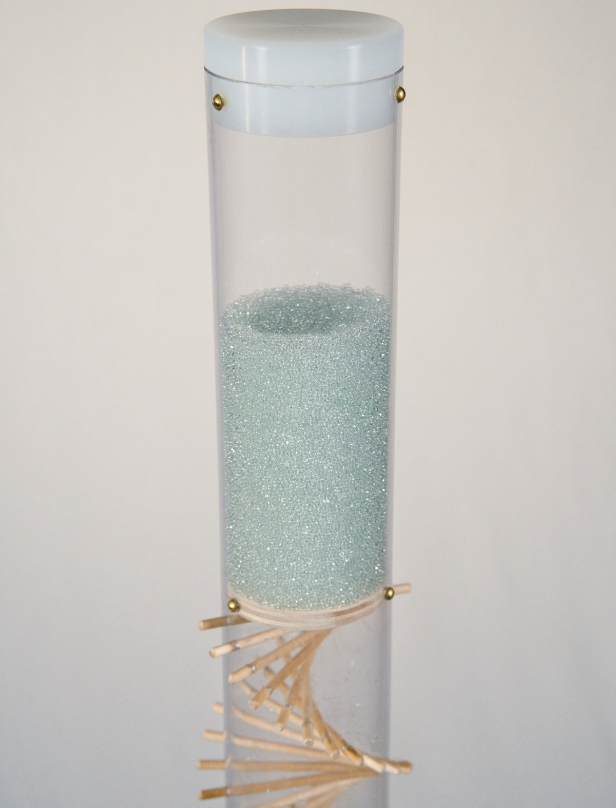 A clear tube contains blue glass microbeads that sparkle, glisten and shine. There is a white cap on top of the rainstick tube. There is a visible wooden spiral running through the clear tube.