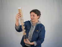 A person holding a 2 foot tall clear rainstick with a wooden spiral going through it. The rainstick is filled with tan amaranth seeds, and the person has a gentle smile on their face. They are calm and relaxed while looking at the rainstick.