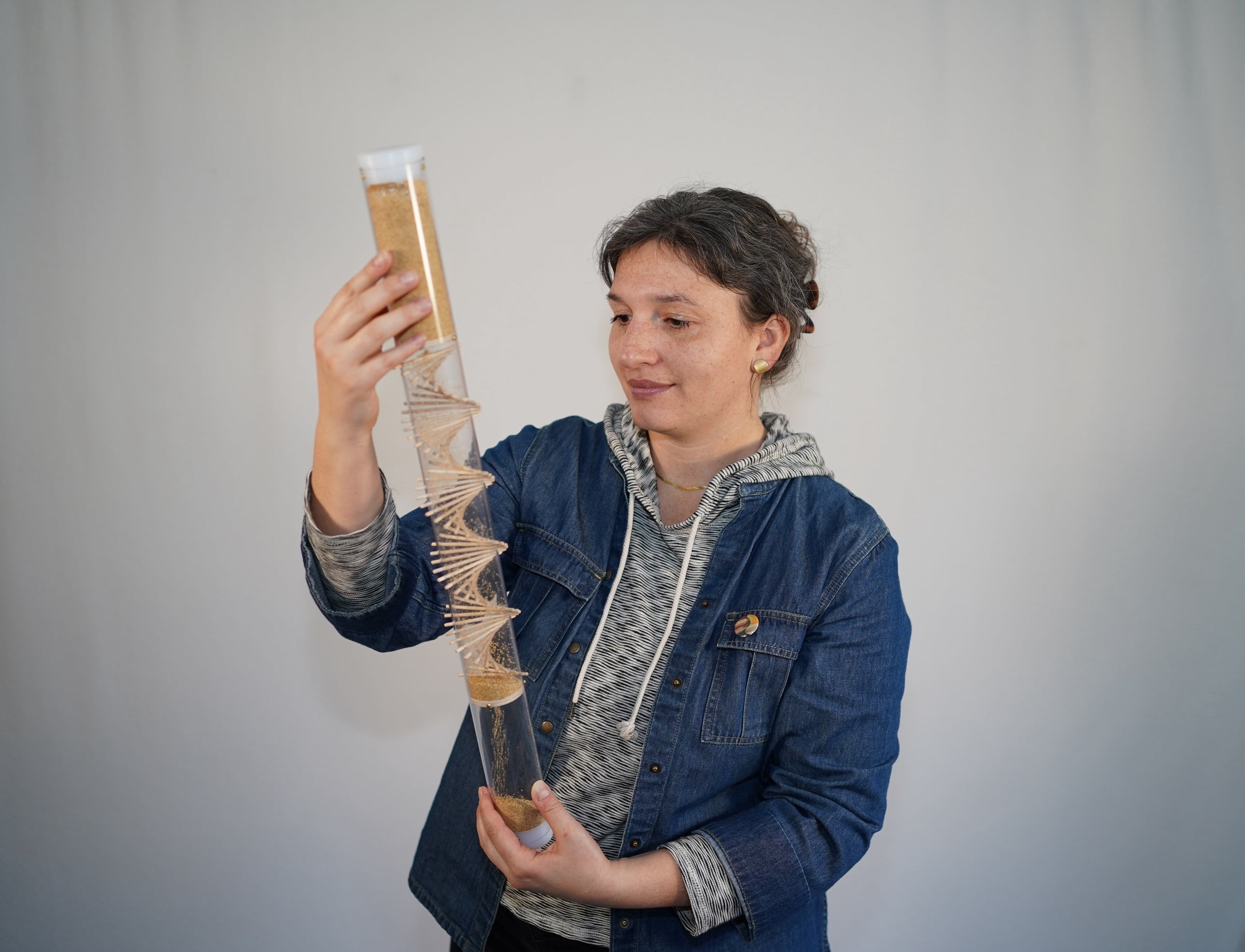A person holding a 2 foot tall clear rainstick with a wooden spiral going through it. The rainstick is filled with tan amaranth seeds, and the person has a gentle smile on their face. They are calm and relaxed while looking at the rainstick.