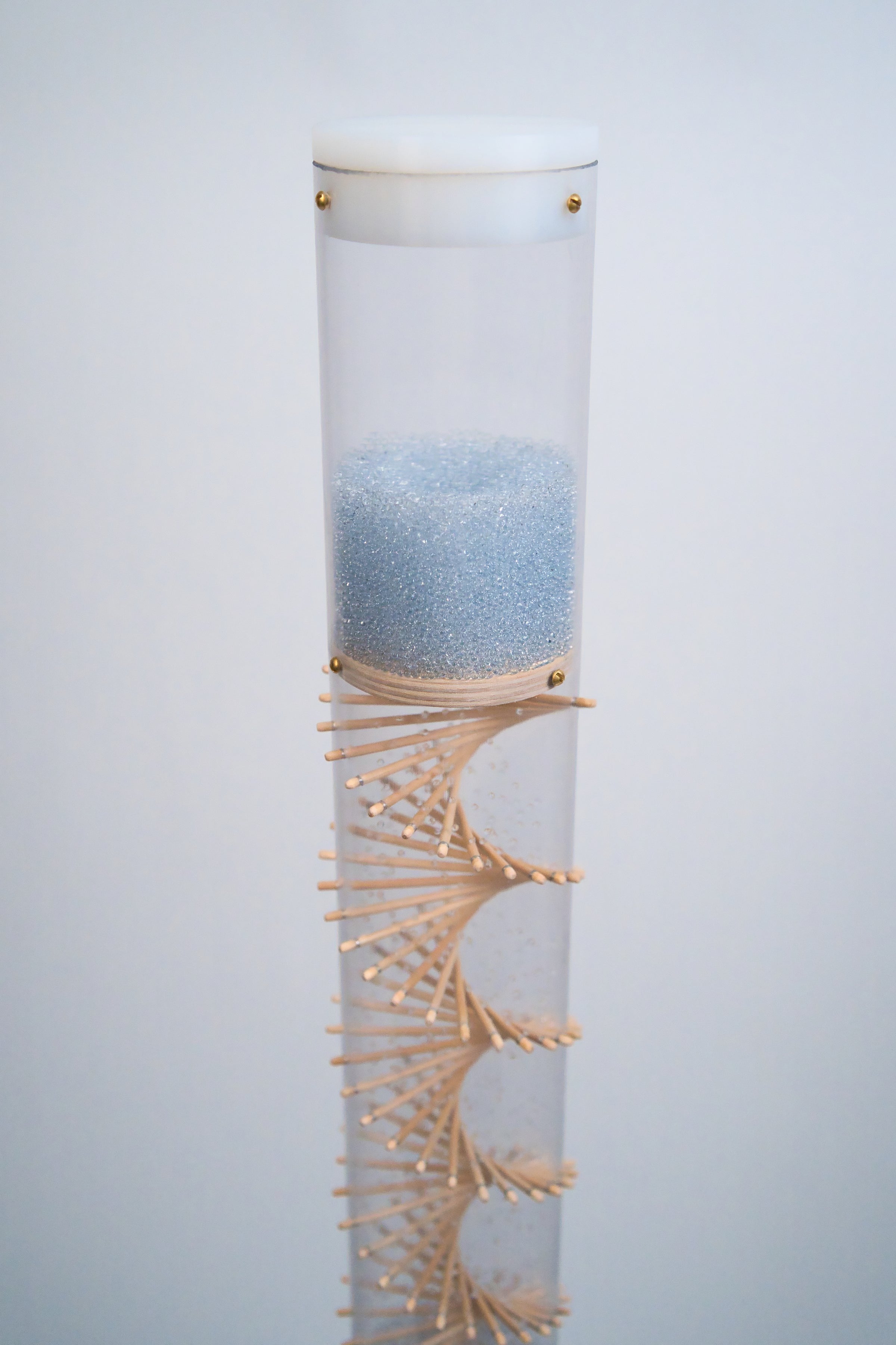 A clear tube with wooden sticks protruding in a spiral pattern is against a white background. The top of the tube is enclosed with a white end cap. Pale blue beads are at the top and flow down to make a sound. This is a long duration rainstick by Rainstick Dreams.