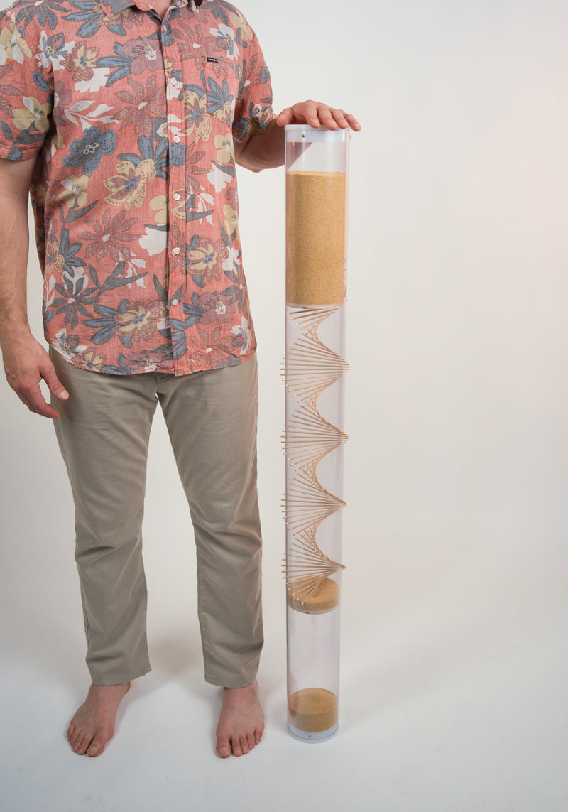 Person standing next to an xl amaranth rainstick by Rainstick Dreams. The person is resting their hand on top of the 20 minute long duration instrument that stands at four feet tall.
