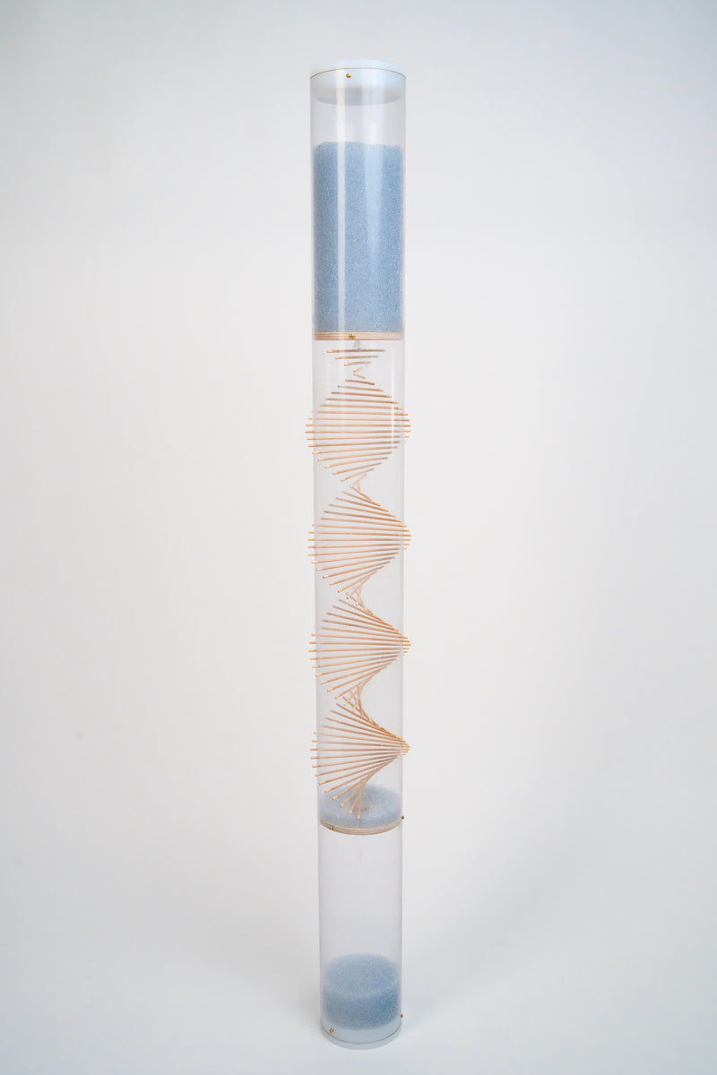 An extra large clear spiral sounds rainstick by Rainstick Dreams is freestanding against a white background. There is a spiral helix comprised of wooden sticks going through the tube. The pale blue glass beads flow down the rainstick. White end caps enclose the top and bottom of the clear tube.