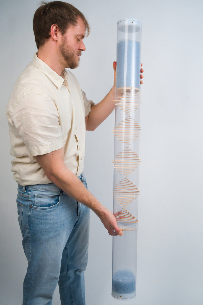 A man wearing a cream button down shirt and pale blue jeans is holding an extra large 4’ tall spiral sounds rainstick in his hands. He is looking at the rainstick and is gently smiling at it. The rainstick has pale blue glass beads flowing from the top, through a wooden spiral helix, down through a funnel disc, into the bottom reservoir of the clear tube. The top and bottom of the clear Rainstick tube and enclosed with white end caps.