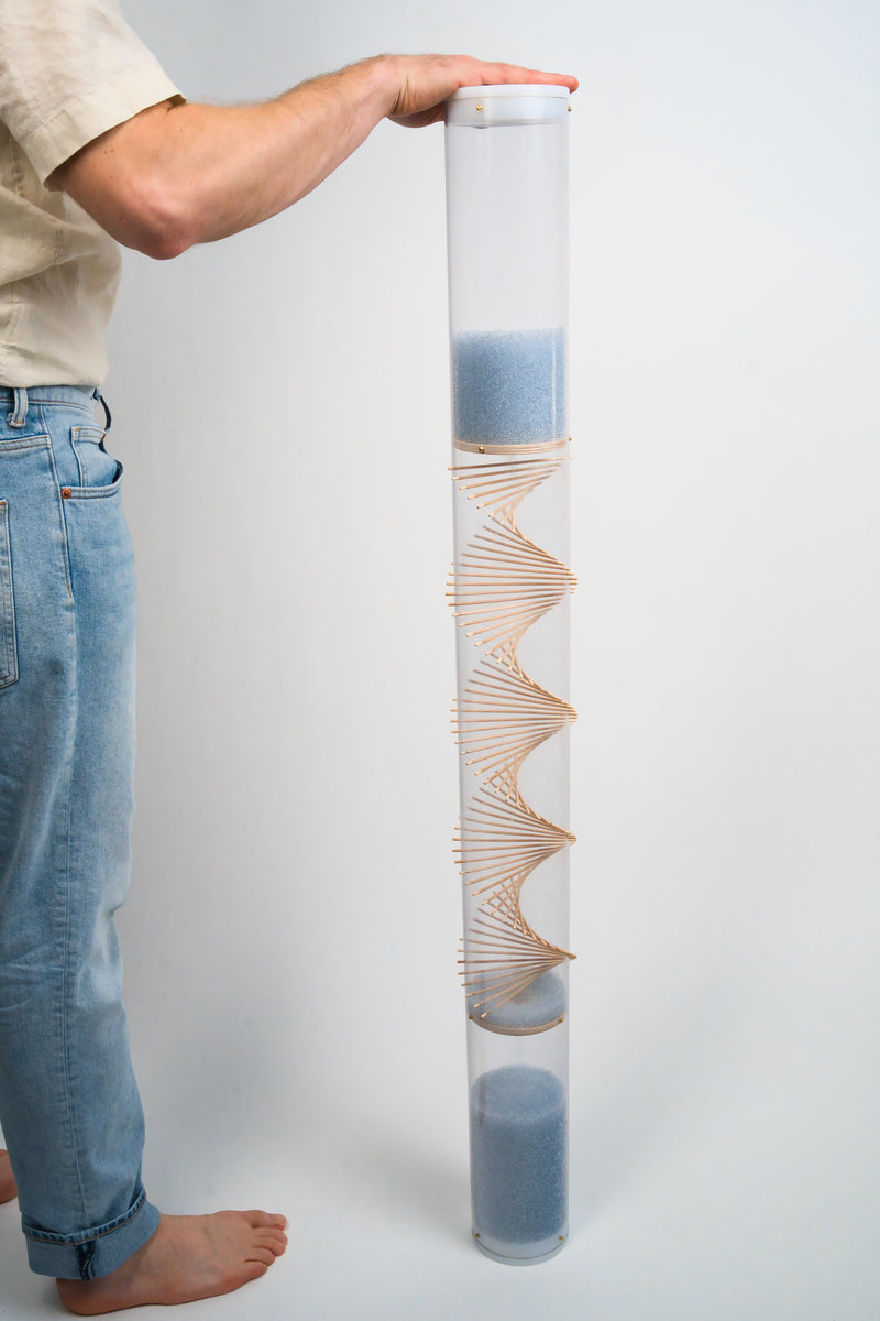 A barefoot person is standing next to an Extra Large 4’ tall spiral sounds rainstick by Rainstick Dreams. Their hand is on the top white end cap that encloses the clear tube. The rainstick is comprised of a clear tube with a wooden spiral helix going through it. The blue glass beads flow from top to bottom. Another white end cap encloses the tube at the bottom.